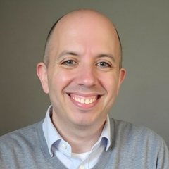 Portrait of Adam S. Dince, smiling, in a gray sweater over a light blue collared shirt.