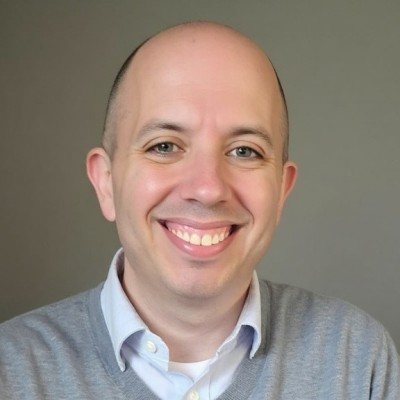 Portrait of Adam S. Dince, smiling, in a gray sweater over a light blue collared shirt.